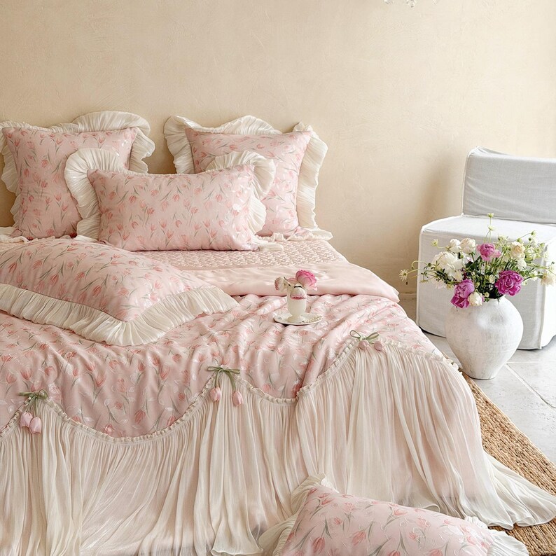 Puede incluir: Un juego de cama de tela rosa suave con un delicado estampado de tulipanes. El edred&oacute;n y las fundas de almohada est&aacute;n adornados con bordes de volantes y detalles de tela plisada en color crema. El conjunto incluye varias almohadas y colcha.