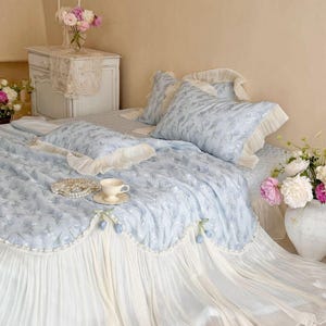 May include: A light blue bedding set featuring a delicate floral pattern, ruffled cream-colored edges, and a pleated skirt. The set includes a quilt, matching pillowcases, and a decorative bolster pillow adorned with small blue tulip accents.