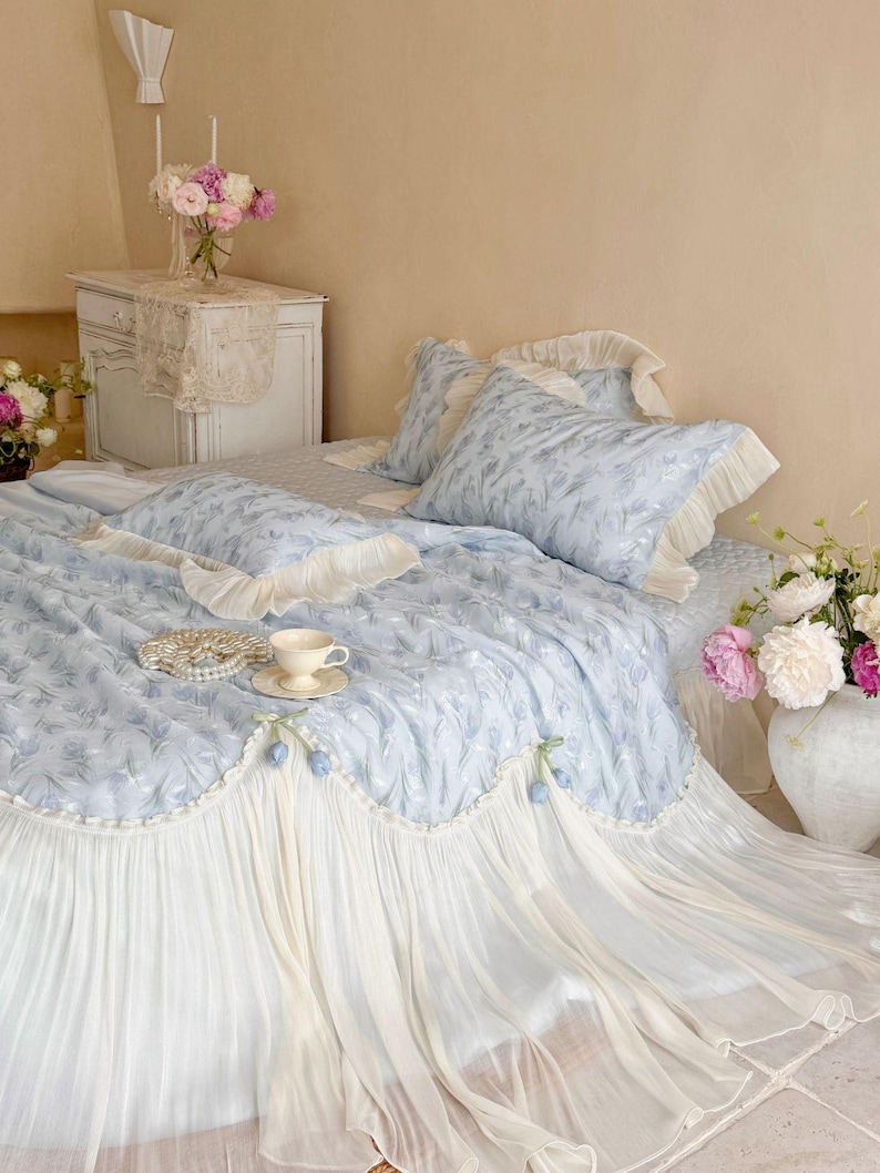 May include: A light blue bedding set featuring a delicate floral pattern, ruffled cream-colored edges, and a pleated skirt. The set includes a quilt, matching pillowcases, and a decorative bolster pillow adorned with small blue tulip accents.