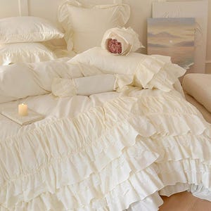 May include: Cream-colored bedding set with ruffled edges and lace detailing. The set includes pillows, a duvet, and a decorative bolster pillow. A lit candle sits on a small book, and a bouquet of pink roses is on the bed.