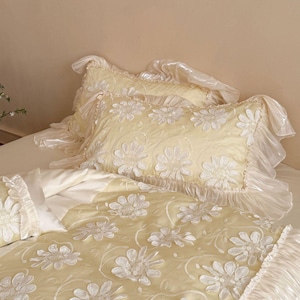 May include: A pale yellow bedding set featuring a duvet cover and two pillowcases with white floral appliqu&eacute; patterns. The edges of the pillowcases and the duvet cover are finished with wide, ruffled, semi-sheer white fabric trim.