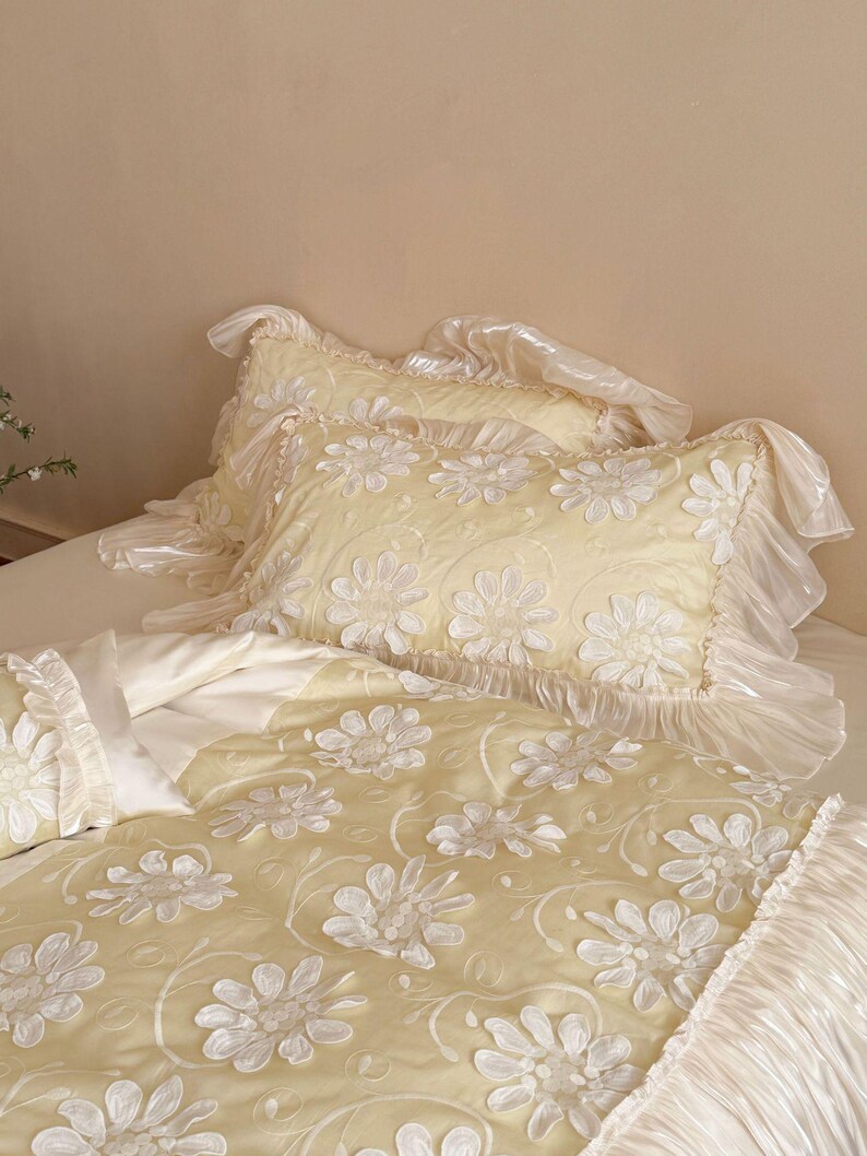 May include: A pale yellow bedding set featuring a duvet cover and two pillowcases with white floral appliqu&eacute; patterns. The edges of the pillowcases and the duvet cover are finished with wide, ruffled, semi-sheer white fabric trim.