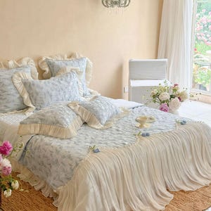 May include: A bed set featuring a light blue and white floral patterned comforter and matching pillows with cream-colored ruffled edges. The comforter includes a long, pleated cream-colored fabric skirt that drapes over the sides of the bed.