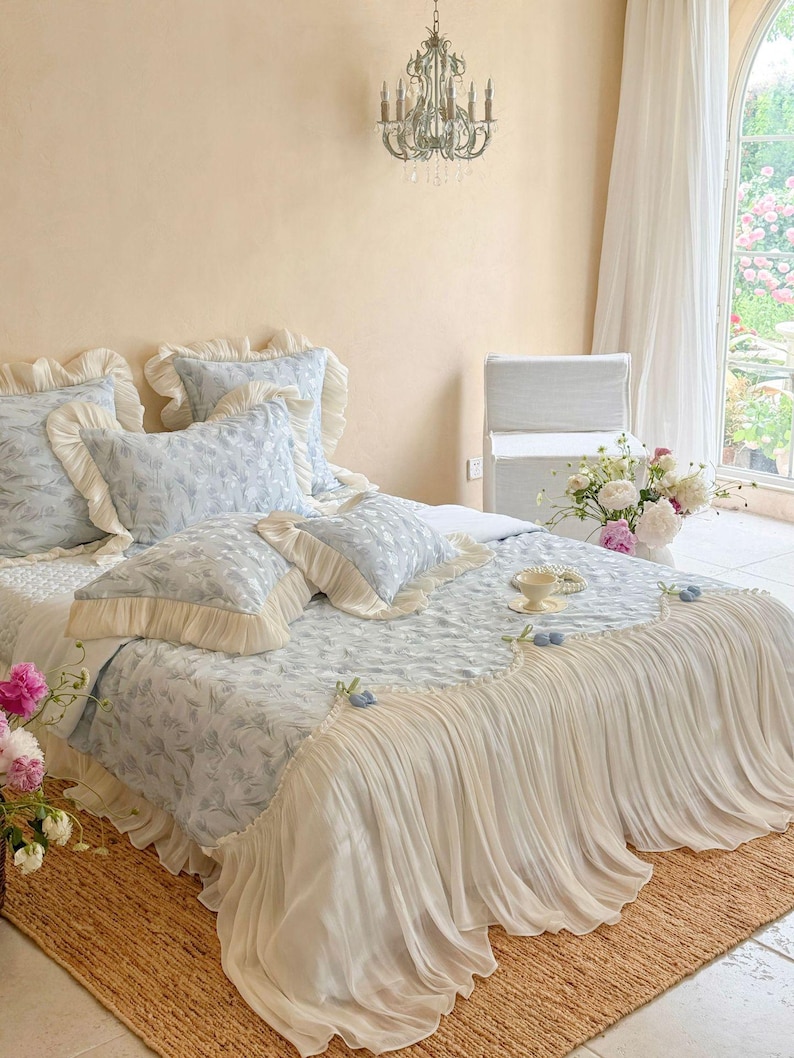 May include: A bed set featuring a light blue and white floral patterned comforter and matching pillows with cream-colored ruffled edges. The comforter includes a long, pleated cream-colored fabric skirt that drapes over the sides of the bed.