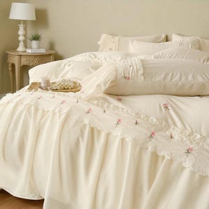 May include: Cream-colored bedding set with embroidered pink floral accents. The set includes a bed skirt with a scalloped lace trim, a duvet cover, and pillows. A side table with a lamp and a small plant is visible in the background.