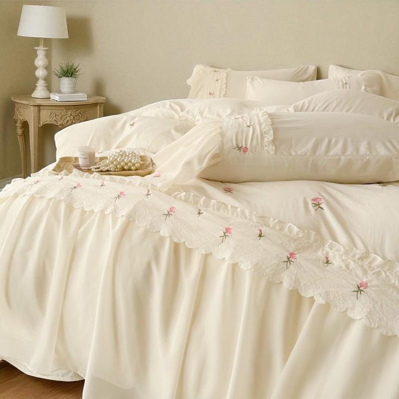 May include: Cream-colored bedding set with embroidered pink floral accents. The set includes a bed skirt with a scalloped lace trim, a duvet cover, and pillows. A side table with a lamp and a small plant is visible in the background.