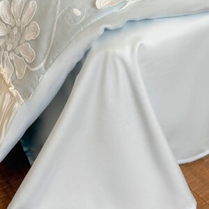 May include: A light blue bedspread featuring large white floral embroidery with intricate petal details and swirling vine patterns. The fabric has a smooth, satin-like finish with a draped corner and a ruffled edge detail along the side.