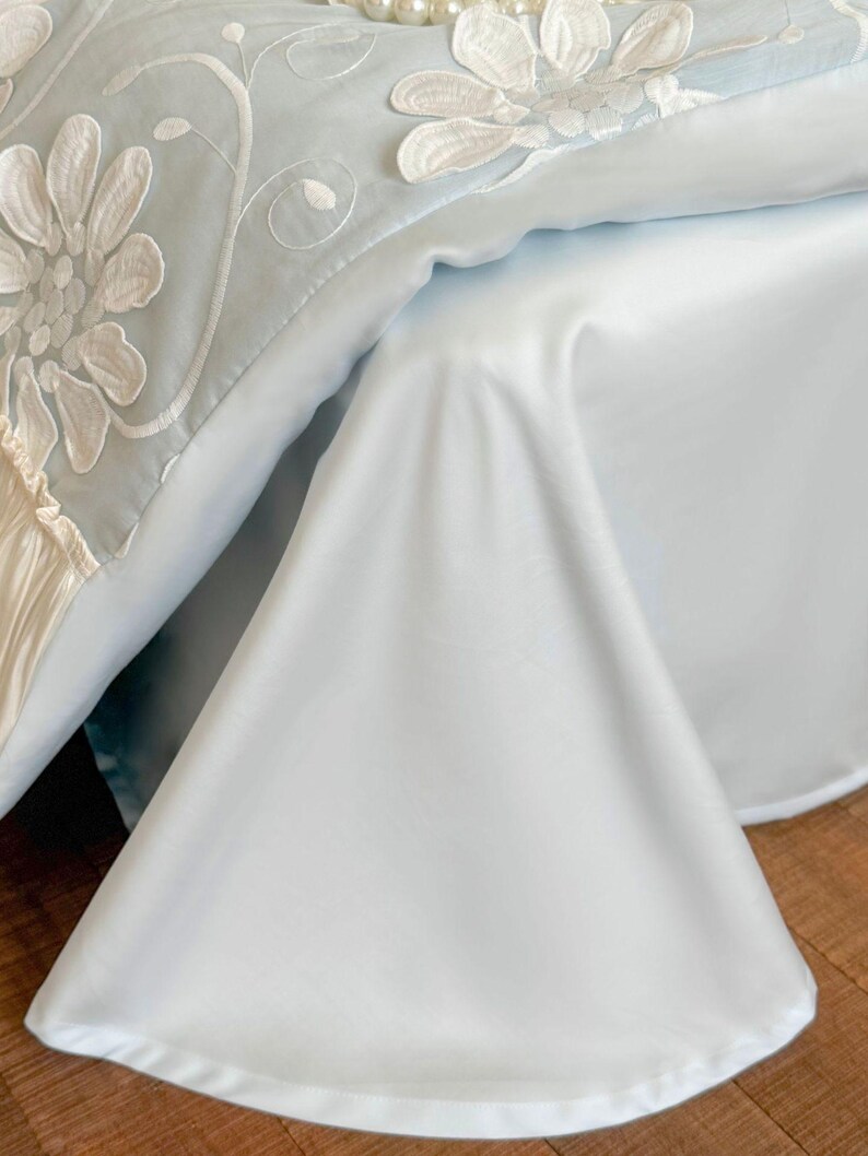 May include: A light blue bedspread featuring large white floral embroidery with intricate petal details and swirling vine patterns. The fabric has a smooth, satin-like finish with a draped corner and a ruffled edge detail along the side.