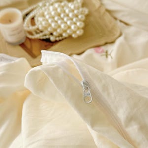 May include: Close-up of a cream-colored pillowcase with a white zipper. A pearl necklace, a lit candle, and a decorative piece are in the background. The bedding has a soft, inviting aesthetic.