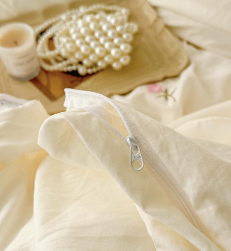 May include: Close-up of a cream-colored pillowcase with a white zipper. A pearl necklace, a lit candle, and a decorative piece are in the background. The bedding has a soft, inviting aesthetic.