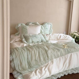 May include: A bedding set featuring a white duvet cover and matching pillowcases with sage green, ruffled, pleated fabric overlays. The set includes decorative bows and small pearl-like accents on the textured green sections, creating a layered look.