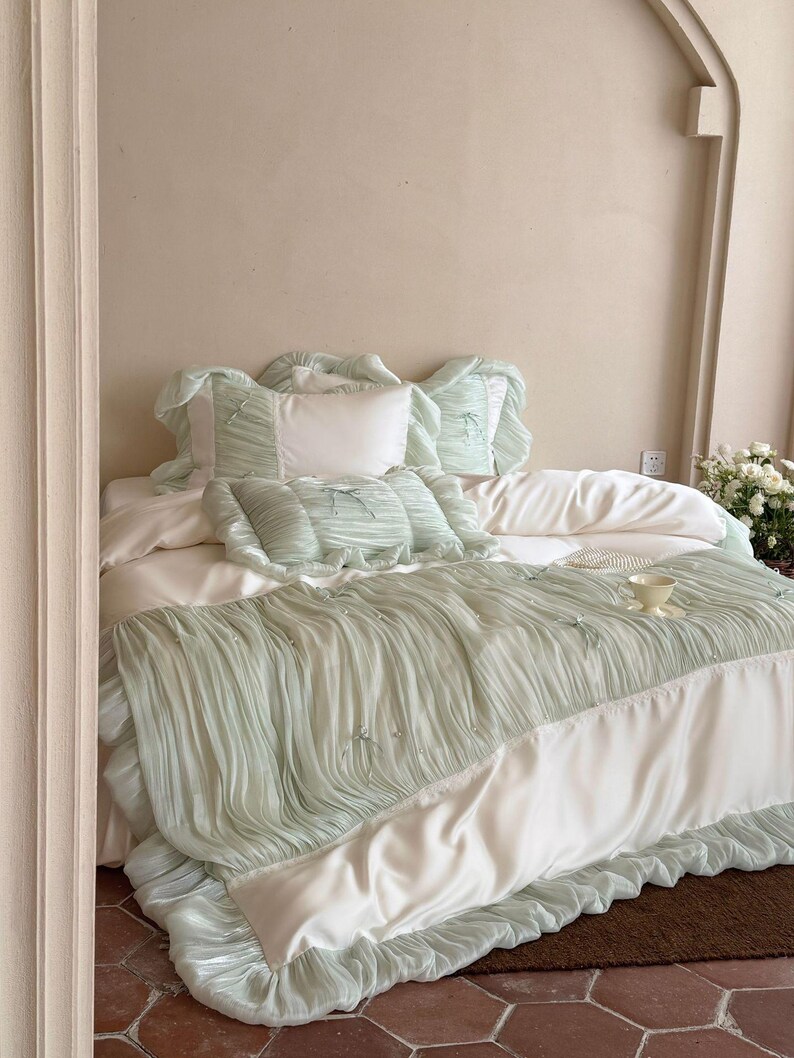 May include: A bedding set featuring a white duvet cover and matching pillowcases with sage green, ruffled, pleated fabric overlays. The set includes decorative bows and small pearl-like accents on the textured green sections, creating a layered look.