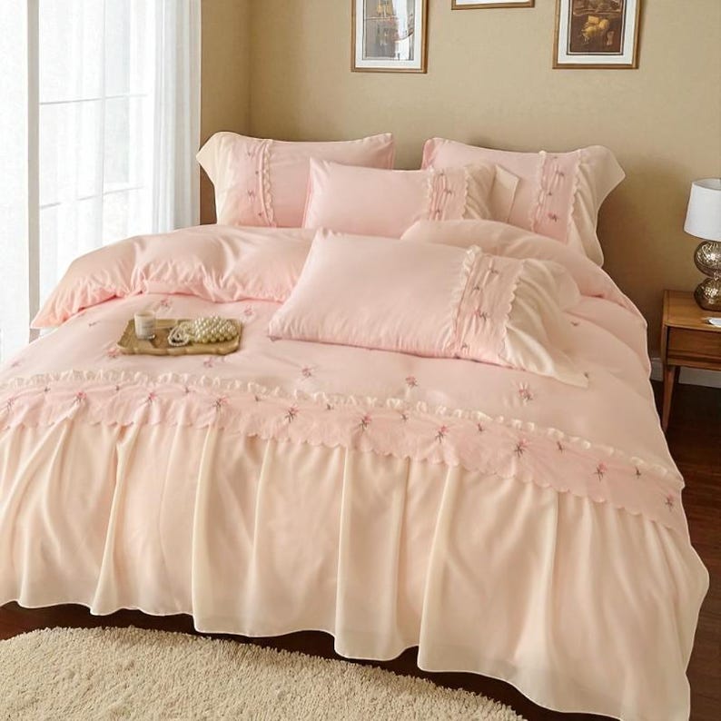 May include: A light pink bed set with multiple pillows and a comforter. The comforter and pillowcases have decorative trim and embroidered floral accents. A wooden tray with a cup and pearls sits on the bed.