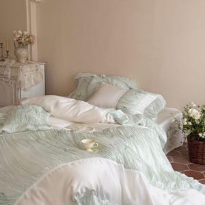 May include: A bedding set featuring a light mint green ruched duvet cover and matching pillowcases with white fabric accents. The set includes a smooth white flat sheet, a small square white pillow, and decorative pearl-like embellishments.