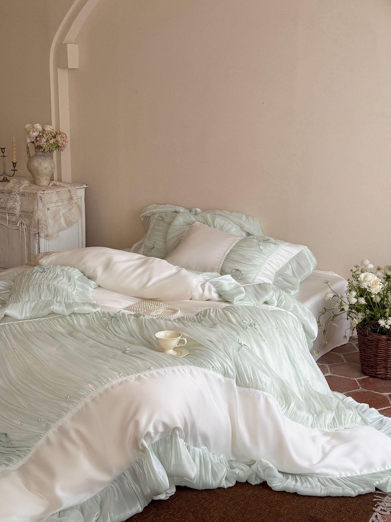 May include: A bedding set featuring a light mint green ruched duvet cover and matching pillowcases with white fabric accents. The set includes a smooth white flat sheet, a small square white pillow, and decorative pearl-like embellishments.