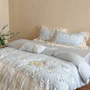 May include: A light blue bedding set featuring a white floral embroidery pattern with raised petals. The duvet cover and pillowcases are accented with ruffled cream-colored fabric borders, creating a layered texture across the bed surface.