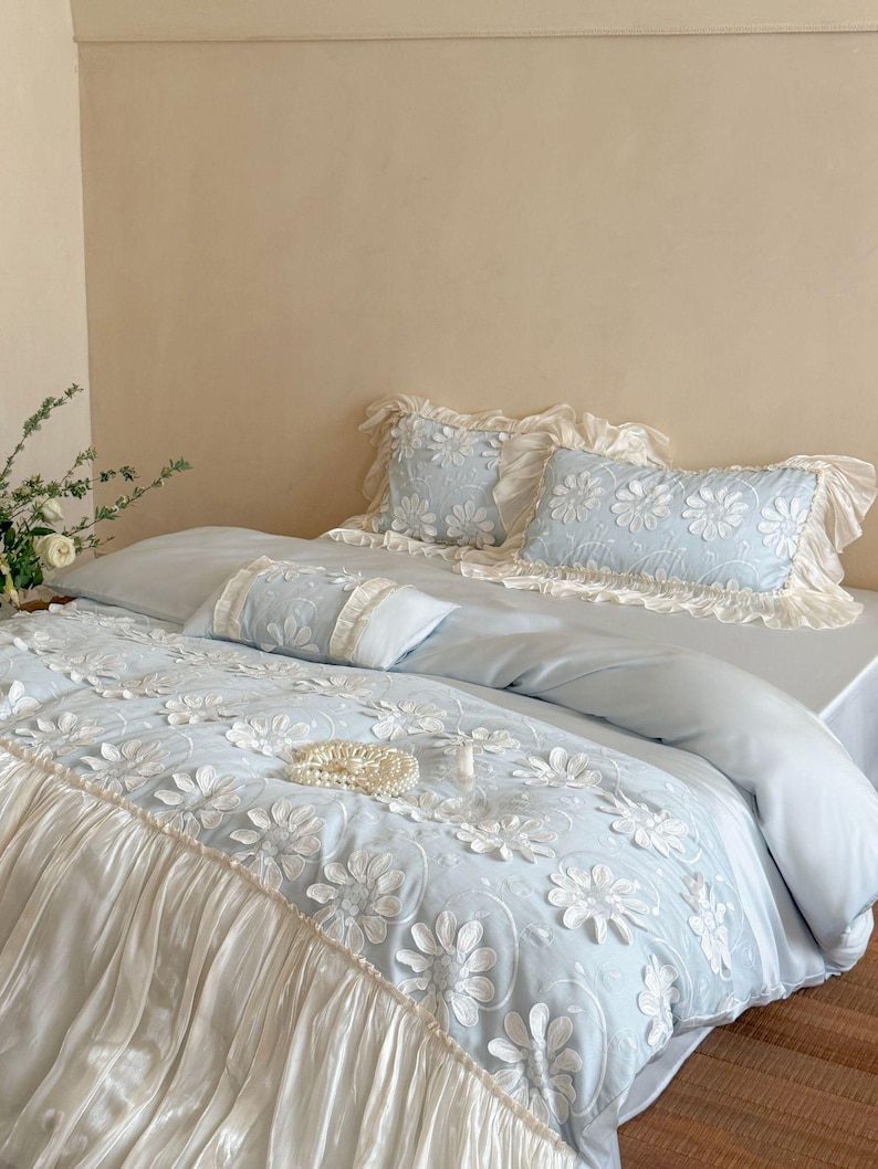 May include: A light blue bedding set featuring a white floral embroidery pattern with raised petals. The duvet cover and pillowcases are accented with ruffled cream-colored fabric borders, creating a layered texture across the bed surface.