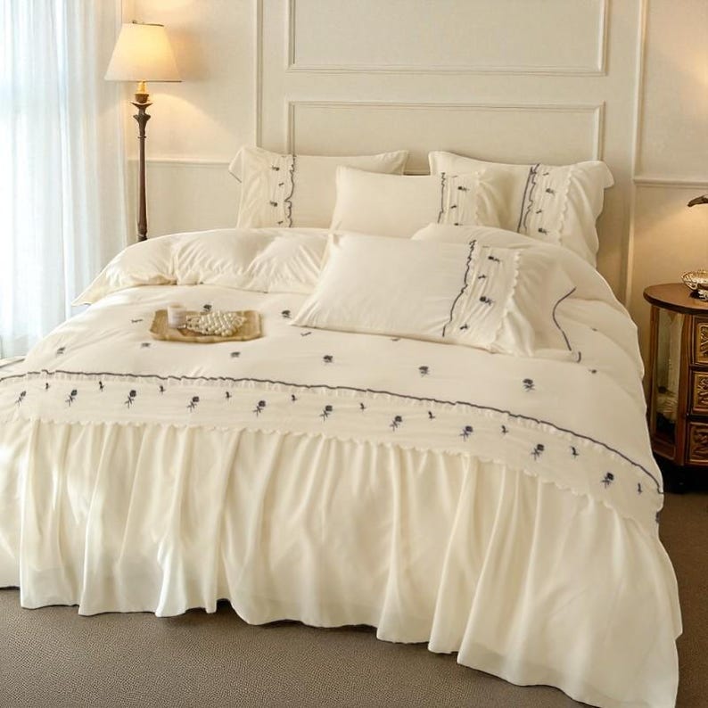 May include: Cream-colored bed linens with a ruffled bed skirt and embroidered floral details. The bedding set includes pillows and a comforter, creating a coordinated and elegant bedroom ensemble.