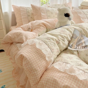 Puede incluir: Una cama con edredón y almohadas con estampado floral. El edredón y las almohadas tienen un patrón de cuadros rosa y blanco con ribete de encaje blanco. Un oso de peluche está en la cama y una revista con la palabra "Chloe" es visible.