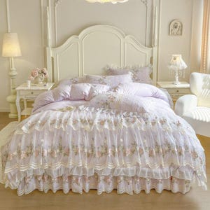 Könnte beinhalten: Ein Bett mit weißem Kopfteil und passenden Nachttischen. Die Bettwäsche ist helllila mit floralen Mustern und einem gerüschten Rock. Die Tagesdecke hat eine zarte Spitzentrimmung mit floraler Stickerei.