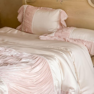 May include: A bedding set featuring a smooth cream-colored duvet cover and matching pillowcases. The set is accented with panels of pleated light pink fabric, delicate ruffles along the edges, and small white pearl-like embellishments.