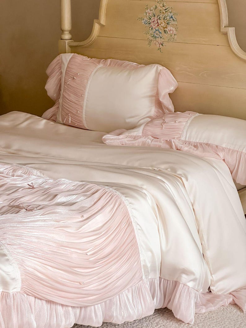 May include: A bedding set featuring a smooth cream-colored duvet cover and matching pillowcases. The set is accented with panels of pleated light pink fabric, delicate ruffles along the edges, and small white pearl-like embellishments.