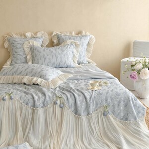 May include: A light blue bedding set featuring a delicate floral pattern, including two square ruffled pillows, one rectangular ruffled pillow, and a bedspread with a wide, pleated cream-colored skirt adorned with small blue flower accents.