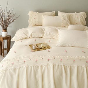 May include: Cream-colored bedding set with embroidered pink roses. The set includes a duvet cover, pillows, and a bed skirt with a scalloped edge. A small tray with a candle and pearls sits on the bed, adding a touch of elegance to the bedroom decor.