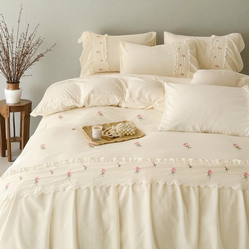May include: Cream-colored bedding set with embroidered pink roses. The set includes a duvet cover, pillows, and a bed skirt with a scalloped edge. A small tray with a candle and pearls sits on the bed, adding a touch of elegance to the bedroom decor.