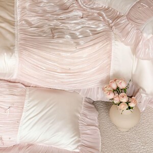 May include: A bedding set featuring a light pink ruched fabric with small pearl accents and thin ribbon ties. The set includes a comforter and matching pillow shams with smooth cream-colored panels and ruffled edges, paired with a vase of pink roses.