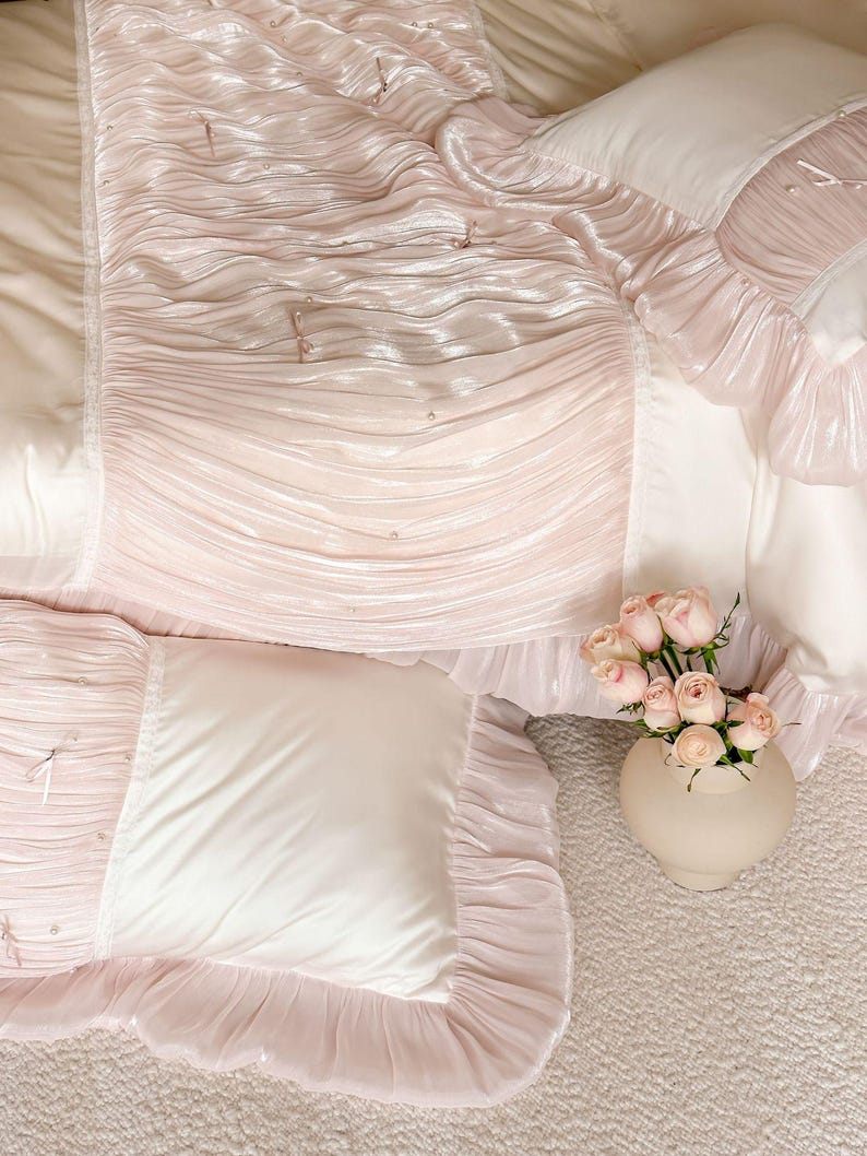 May include: A bedding set featuring a light pink ruched fabric with small pearl accents and thin ribbon ties. The set includes a comforter and matching pillow shams with smooth cream-colored panels and ruffled edges, paired with a vase of pink roses.