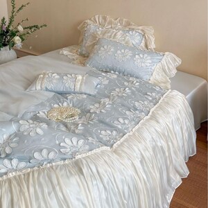 May include: A light blue bedding set featuring white embroidered daisy patterns, including a duvet cover, two rectangular pillows, and a smaller accent pillow. The set is finished with ruffled cream-colored fabric trim along the edges.