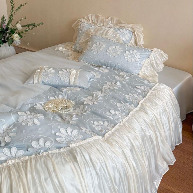 May include: A light blue bedding set featuring white embroidered daisy patterns, including a duvet cover, two rectangular pillows, and a smaller accent pillow. The set is finished with ruffled cream-colored fabric trim along the edges.