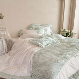 May include: A bedding set featuring a white flat sheet paired with a light mint green ruched comforter. The comforter is decorated with small white pearls and thin ribbon bows. The set includes matching ruffled pillow shams and a white pillow.