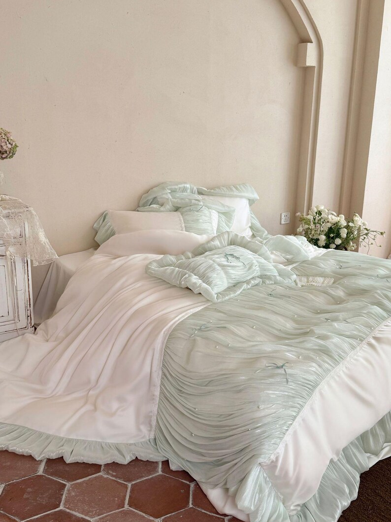 May include: A bedding set featuring a white flat sheet paired with a light mint green ruched comforter. The comforter is decorated with small white pearls and thin ribbon bows. The set includes matching ruffled pillow shams and a white pillow.