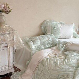 May include: A bedding set featuring a light mint green duvet cover and matching pillowcases with a ruched, pleated texture. The set includes smooth white fabric panels, small decorative bows, and a small white beaded handbag resting on top.