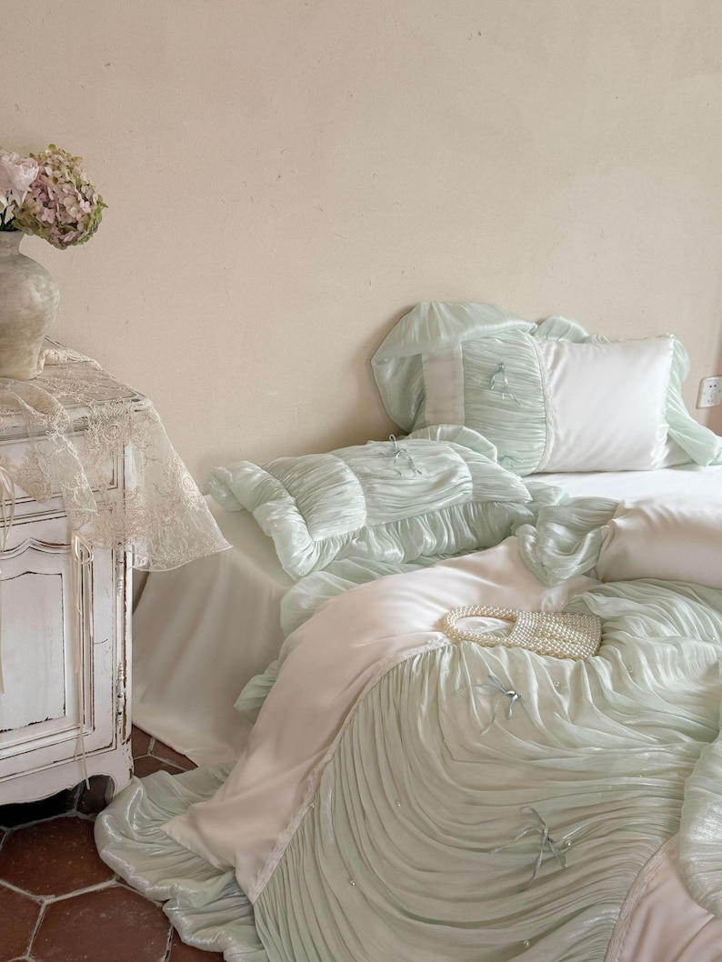 May include: A bedding set featuring a light mint green duvet cover and matching pillowcases with a ruched, pleated texture. The set includes smooth white fabric panels, small decorative bows, and a small white beaded handbag resting on top.