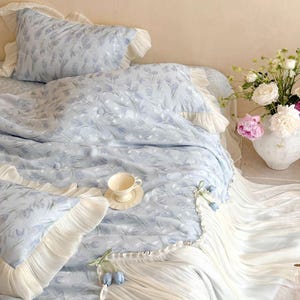 May include: A light blue bedding set featuring a repeating tulip pattern, accented with cream-colored ruffled edges and decorative fabric tulip buds. The set includes a duvet cover and matching pillowcases, all finished with pleated fabric trim.