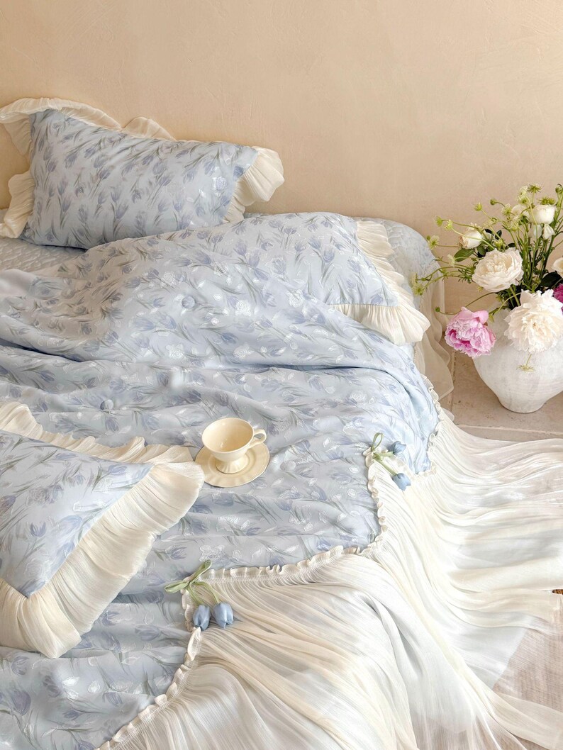 May include: A light blue bedding set featuring a repeating tulip pattern, accented with cream-colored ruffled edges and decorative fabric tulip buds. The set includes a duvet cover and matching pillowcases, all finished with pleated fabric trim.
