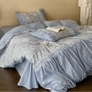 Peut inclure: Un ensemble de literie bleu clair avec des motifs floraux blancs et des bordures en dentelle. L'ensemble comprend une housse de couette, des taies d'oreiller et un coussin décoratif. Un livre ouvert repose sur les oreillers.