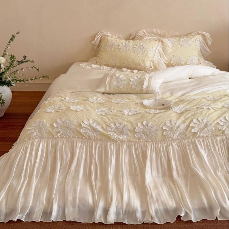 May include: A pale yellow bedding set featuring a duvet cover and matching pillowcases adorned with white, three-dimensional floral appliqu&eacute;s. The duvet cover includes a wide, gathered, solid cream-coloured fabric ruffle along the bottom edge.