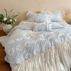 May include: A light blue bedding set featuring white floral applique patterns and ruffled edges. The set includes a duvet cover with a large floral design, two rectangular pillows with ruffled borders, and a smaller rectangular accent pillow.