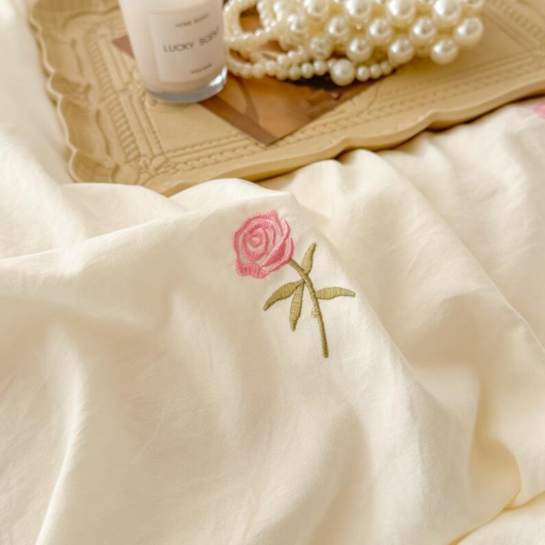 May include: Cream-colored bedding with a delicate embroidered pink rose and green stem. A candle and a string of pearls rest on a decorative tray in the background. The bedding has a soft, textured appearance.