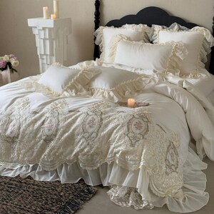 May include: A luxurious white bed set with decorative lace and floral embroidery. The bedding includes pillows with ruffled edges, a comforter, and a bed skirt. A black bed frame and candles complete the elegant bedroom scene.