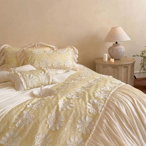 May include: A bedding set featuring a pale yellow runner and matching pillow shams adorned with white floral appliqu&eacute;s. The runner has a pleated cream-coloured skirt, and the pillows are finished with ruffled edges, creating a layered look.