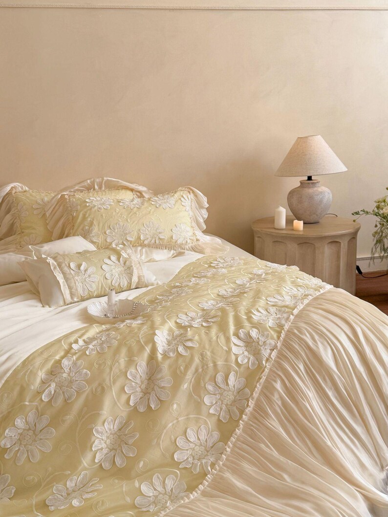 May include: A bedding set featuring a pale yellow runner and matching pillow shams adorned with white floral appliqu&eacute;s. The runner has a pleated cream-coloured skirt, and the pillows are finished with ruffled edges, creating a layered look.