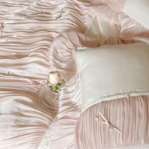 May include: A light pink bedding set featuring a ruffled, pleated texture with small white pearl accents and thin ribbon ties. The set includes a matching pillowcase with a smooth white panel, lace trim, and a single pink rose in a glass.