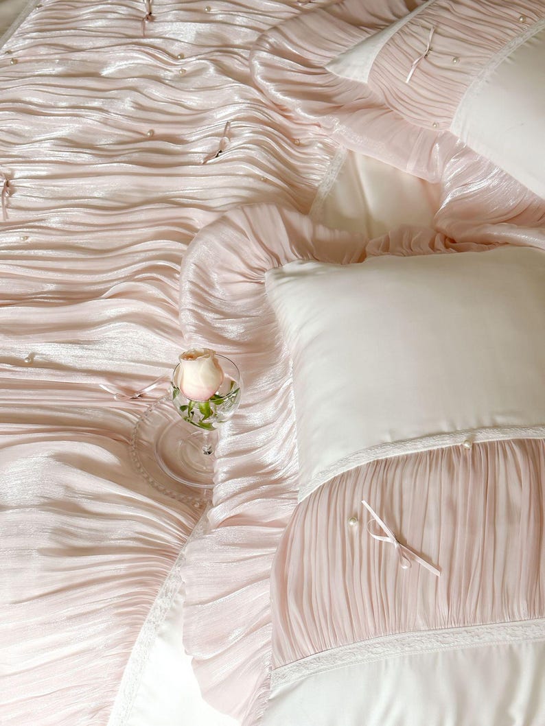 May include: A light pink bedding set featuring a ruffled, pleated texture with small white pearl accents and thin ribbon ties. The set includes a matching pillowcase with a smooth white panel, lace trim, and a single pink rose in a glass.