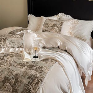 May include: A luxurious bed set with a cream-coloured duvet and pillowcases with ruffled edges. The duvet cover and accent pillows feature a toile pattern in grey and brown. A glass of champagne and an open book rest on the bed.