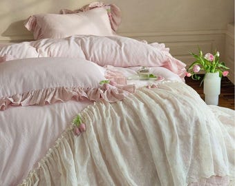 May include: A bed dressed in pink bedding, featuring a ruffled duvet cover and pillowcases. The bed is covered with a white, lace-like bedspread. A vase of pink tulips stands on a wooden floor beside the bed.